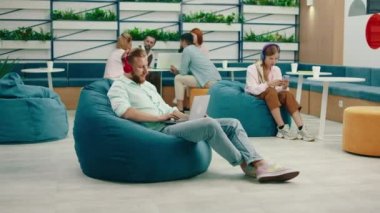 Sarışın sakallı bir işadamı turkuaz fasulye sandalyesinde oturuyor ve kırmızı kablosuz kulaklığıyla müziği dinlerken, arkasındaki kadın da bilgisayarında çalışıyor.