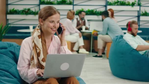 Eine sehr schöne blonde Frau sitzt auf einem blauen Bohnensack und arbeitet an ihrem Laptop, während sie freudig am Telefon spricht. — Stockvideo