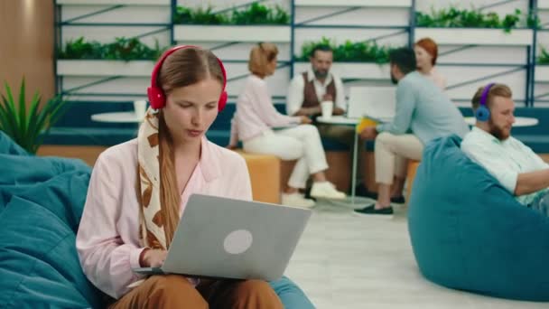 En una sala de oficina con sillas de bolsas de frijoles, un pequeño grupo de personas está celebrando una reunión mientras un hombre lleva auriculares inalámbricos azules y va en su teléfono y una mujer está escuchando música, vibing — Vídeo de stock