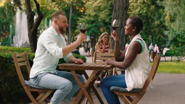 Um jovem homem encantador com cabelo loiro e barba está compartilhando um pouco de vinho com uma bela mulher africana em uma mesa em um parque muito cênico — Vídeo de Stock