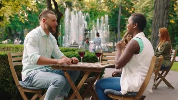 Una mujer africana de buen aspecto con el pelo corto y un hombre blanco rubio con barba se ríen mientras se sientan en una mesa frente a frente en un parque, disfrutando de la compañía de los demás — Vídeos de Stock