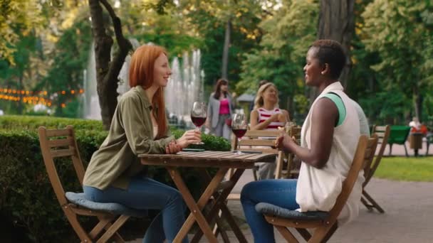 Due donne dall'aspetto coinvolgente sono a un tavolo all'aperto circondate da alberi e natura, a condividere del vino e a conversare — Video Stock