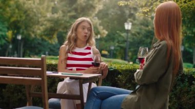 Kızıl saçlı arkadaşıyla, estetik görünümlü parktaki ahşap bir masada oturan ve konuşan çarpıcı sarışın bir kadının düşük açılı görüntüsü.
