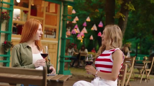 Две очаровательные подруги счастливы быть в компании друг друга, когда они разговаривают и смеются над красивым цветочным кафе в парке, они пьют вино за столом — стоковое видео