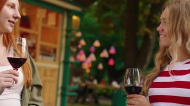 Parkta, sarışın arkadaşıyla bir masada oturan, sohbet eden ve şarap kadehlerini alkışlayan kızıl saçlı kızın yakın plan fotoğrafı. 4k