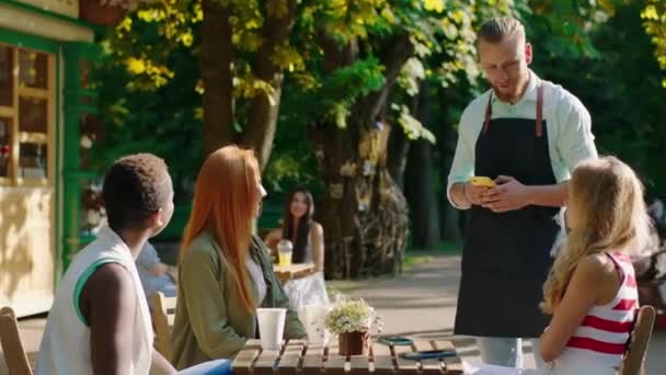 I midten af parken karismatisk barista mand fra cafe tage ordren form en gruppe af fantastiske multiraciale damer med et stort smil – Stock-video