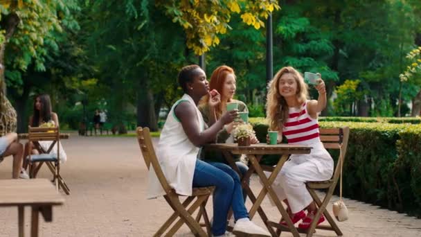 Πολυεθνική καλύτεροι φίλοι κυρίες συνάντηση στο καφέ στη μέση του πάρκου και να τραβήξετε φωτογραφίες με smartphone, ενώ πίνοντας καφέ — Αρχείο Βίντεο