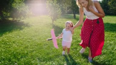 Mutlu ve heyecanlı bir anne ve küçük kızı ellerinde bir uçakla çimlerin üzerinde koşuyorlar. ARRI Alexa Mini 'de çekilen.