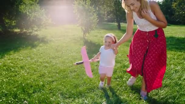 Glückliche und aufgeregte Mama und ihre kleine Tochter rennen vor der Kamera durch das Gras, mit einem Flugzeug in der Hand. Schuss auf ARRI Alexa Mini. — Stockvideo