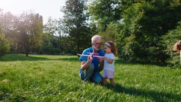 Delante de la cámara en el parque abuelo guapo enseñando a jugar con el avión su nieta linda otro niño pequeño lindo corriendo alrededor — Vídeo de stock