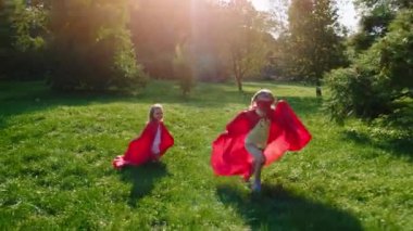 Güneşli bir günde, parkın ortasında iki tatlı çocuk ve süper kahraman kostümü giymiş kız ve erkek çocuk parkın ortasındaki çimlerin arasında hızla koşuyorlar.