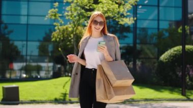 Modern şehrin ortasında, elinde alışveriş poşetleri olan kızıl saçlı kadın kameranın önünde yürüyor ve akıllı telefonuna bir şeyler yazıyor.