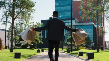 Güneşli bir günde kameranın önünde elinde alışveriş torbalarıyla modern yüksek binadaki siyahi adama doğru yürürken mutlu ve heyecanlı bir şekilde ofisteki iş yerine geri dönüyor. 4k