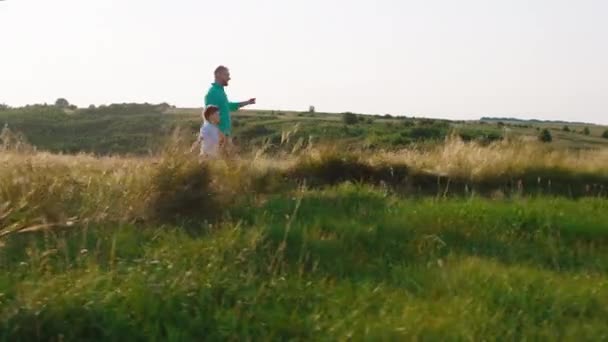 Happy cute and attractive boy and his dad running through the large green field in the middle of nature in a perfect sunny day — Stock Video