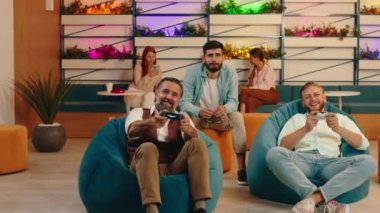 Yaşlı bir adam genç bir adamla bilgisayar oyunu oynuyor, kontrolörleri kullanıyor, arkasındaki adam yaşlı adama tezahürat yapıyor ve hepsini fasulye çuvalında otururken kazanıyor.