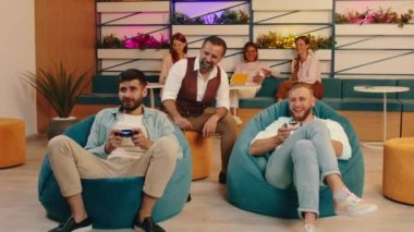 Mavi fasulye koltuklarında oturan iki adam video oyunları oynarken tezahürat yapıyor ve arkalarından yaşlı bir adam onların oynamasını izliyor ayrıca arkada aralarında sohbet eden üç kadın var.