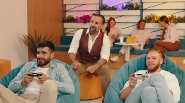 İki erkek video oyunu oynarken birbirleriyle yarışıyor, başka bir yaşlı adam iki adamın arkasında otururken onları izliyor ve arkada üç kadın bir kanepede oturuyor.