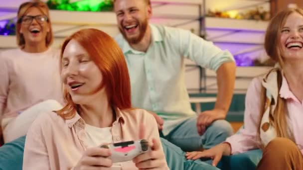 Video oyunlarında kazanan kızıl saçlı bir kadının yakın plan çekimi. Çevresindeki beş kişi onun oynayışını ve mavi fasulye koltuklarında oturuşunu izliyor. — Stok video