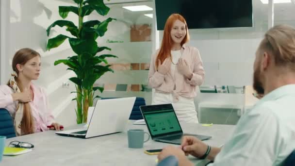 Высокая красивая рыжеволосая женщина стоит в верхней части стола, когда она разговаривает во время встречи с тремя другими людьми в офисе, и она выглядит очень уверенно — стоковое видео