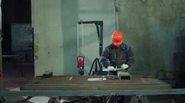 Demirci atölyesinde çekiç ve ağır makineyle çalışıyor. Güvenlik kaskı ve gözlük takıyor..