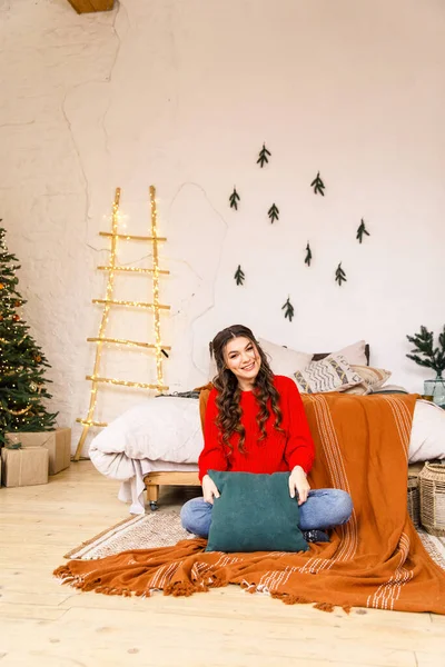 赤いセーターと青いジーンズの若い美しい女の子はクリスマスの装飾で飾られた居心地の良い部屋のベッドの上に座っています — ストック写真