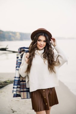 Beyaz kazaklı, kahverengi etekli ve kahverengi şapkalı genç güzel kız gölün kıyısında yürüyor.