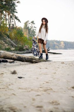 Beyaz kazaklı, kahverengi etekli ve kahverengi şapkalı genç güzel kız gölün kıyısında yürüyor.