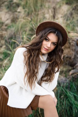 Beyaz kazaklı, kahverengi etekli ve kahverengi şapkalı genç güzel kız gölün kıyısında yürüyor.