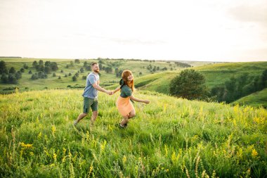 Genç, güzel, kırmızı saçlı, pembe elbiseli, yeşil ceketli, gri tişörtlü, yeşil şortlu bir adam gün batımında çimlerin üzerinde eğleniyor.
