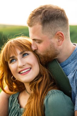 Genç, güzel, kırmızı saçlı, pembe elbiseli, yeşil ceketli, gri tişörtlü, yeşil şortlu bir adam gün batımında çimlerin üzerinde eğleniyor.