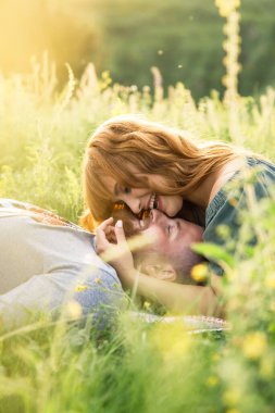 Genç, güzel, kırmızı saçlı, pembe elbiseli, yeşil ceketli, gri tişörtlü, yeşil şortlu bir adam gün batımında çimlerin üzerinde eğleniyor.