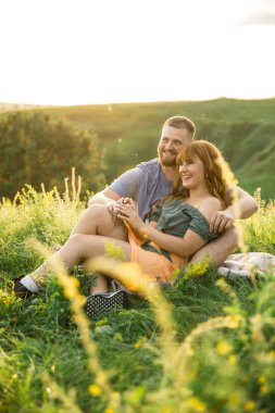 Genç, güzel, kırmızı saçlı, pembe elbiseli, yeşil ceketli, gri tişörtlü, yeşil şortlu bir adam gün batımında çimlerin üzerinde eğleniyor.