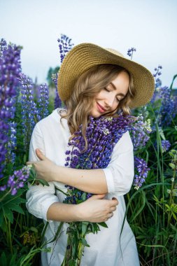 a young beautiful girl in a white dress and hat walks across the field with lupins