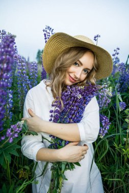 a young beautiful girl in a white dress and hat walks across the field with lupins