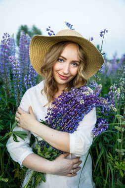 a young beautiful girl in a white dress and hat walks across the field with lupins