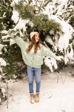 Yeşil kazaklı ve pembe şapkalı genç bir kız kışın ormanda yürüyor.