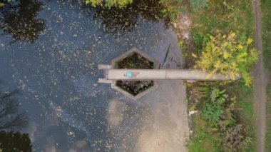 Hava manzaralı, turkuaz kapüşonlu bir adam sonbahar parkında, sarı ağaçlı bir gölün kenarında yatıyor.