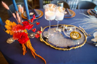 Şölen masası tabaklar, çatal bıçaklar, bardaklar, mumlar ve çiçek düzenlemeleri ile süslenir.