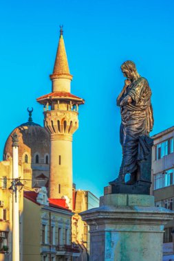 CONSTANTA, ROMANIA-JULY 15, 2020: The statue of Publius Ovidius Naso who was a classical Latin poet,along with Horatiu and Virgil. In the background is the Great Mosque,also known as the Carol Mosque. clipart