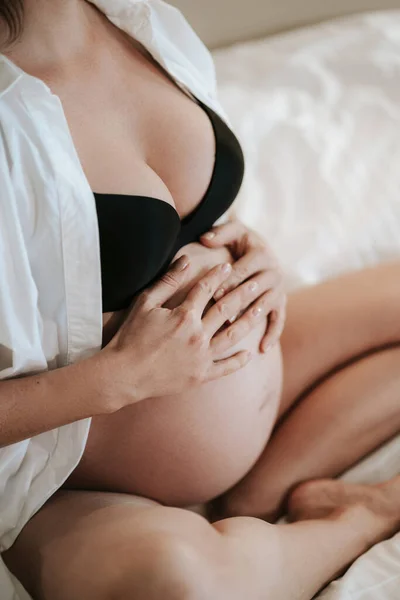 Pregnant woman sits on bed in black bra holding hands on belly