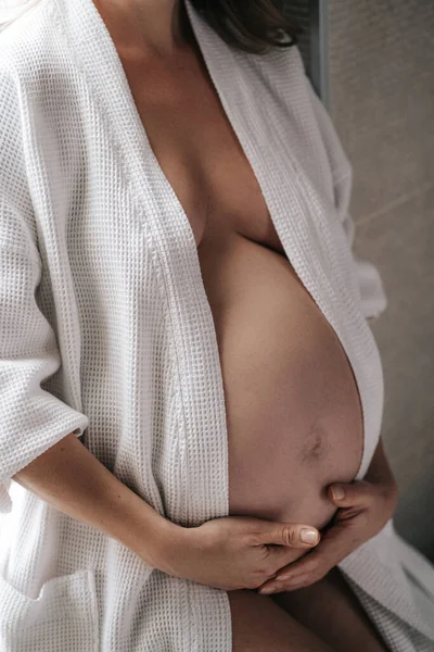 Egy terhes nő fehér kabátban ül, fogja a hasát a kezével. Stock Kép