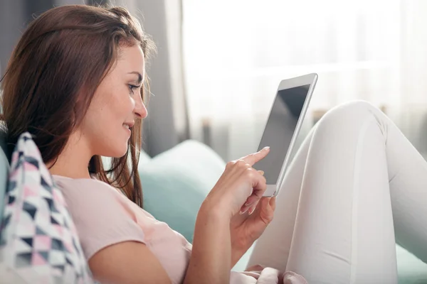 Молодая женщина сидит дома на диване и читает электронную книгу на планшетном компьютере. Стрельба в помещении — стоковое фото