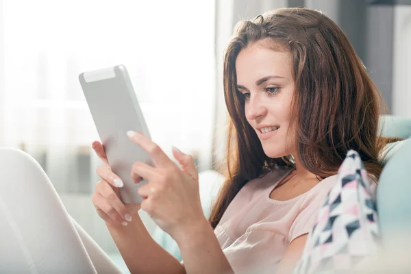 Jovem mulher sentada no sofá em casa e lendo ebook no computador tablet. Casual estilo interior atirar — Fotografia de Stock