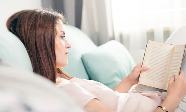 Young woman lying on couch and reading a book at home. Casual style indoor shoot — Stock Fotó