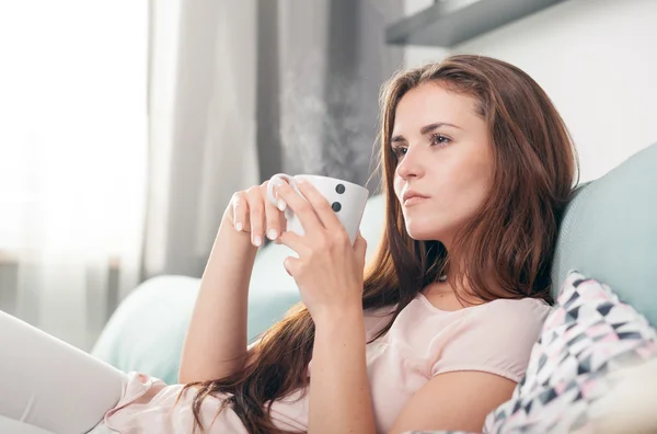 Thinking woman sitting on couch at home and drinking coffee. Casual style indoor shoot