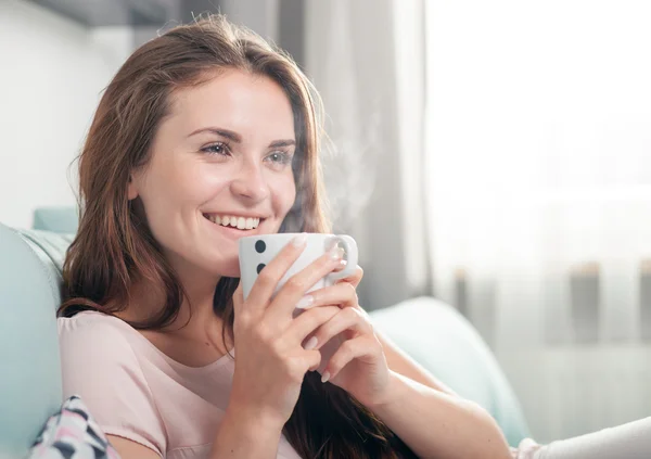 Donna sorridente seduta sul divano a casa e che beve caffè. Scatto al coperto stile casual — Foto Stock