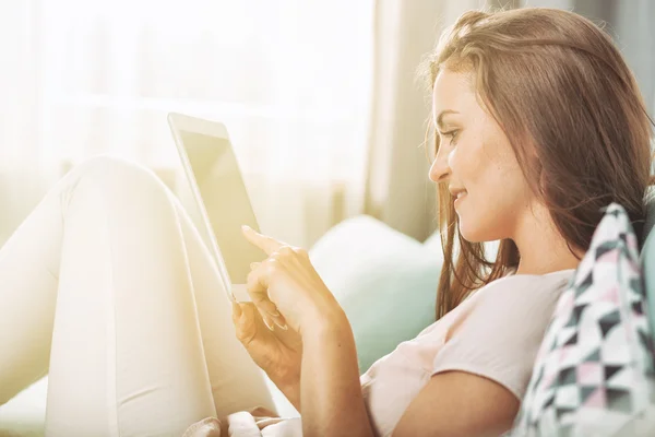 Молодая женщина сидит на диване дома и читает электронную книгу на планшете — стоковое фото