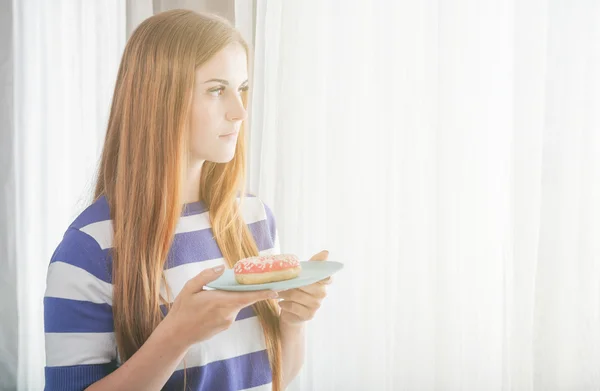 Woman at home looking in the window and holding donat — Stock Photo, Image