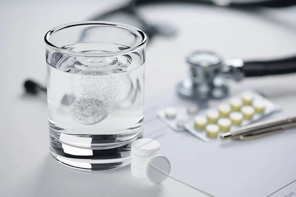 Effervescent tablet in a glass of water, stethoscope and pills on the doctors table.
