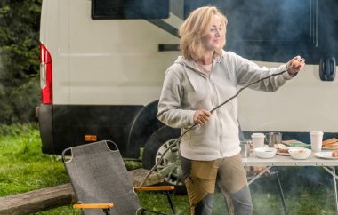 Caucasian Woman in Her 40s Having Fun on a Camping. Preparing Polish Sausage on Her Self Made Cooking Stick. Modern Class B Motorhome in Background.  clipart